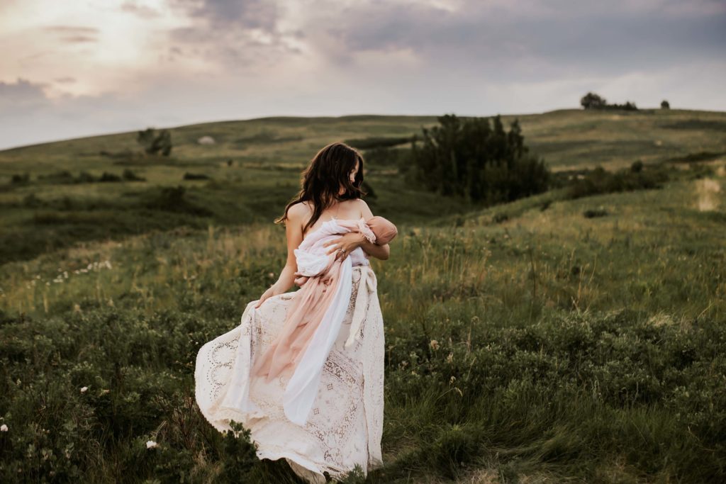 Newborn Photographer, a mother holds her baby while walking on a grassy hillside. She wears a white dress
